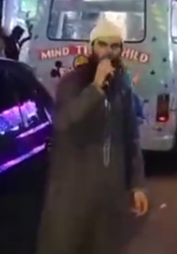 A still of Abu Ibraheem Hussnayn, a young South Asian man wearing a white cap and with a long beard wearing a long, dark-coloured robe, talking into a microphone he is holding in his hand, standing in front of an ice-cream van with the slogan "Mind that child" on it.