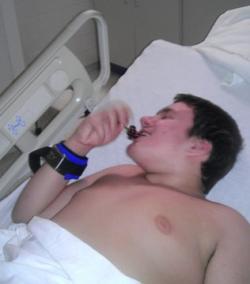 Picture of Alex Spourdalakis, a white teenage boy, lying in a hospital bed with a cuff around his right wrist which is attached to the bed, with him holding an object of some kind in his mouth. He is covered by a white sheet from his waist down.