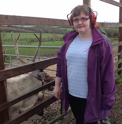 Picture of Claire Dyer, a young white woman wearing glasses, red sound blockers, a light blue and white striped T-shirt and a purple jacket, standing next to a fence behind which is a male deer