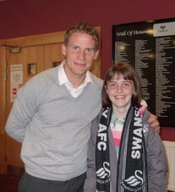 Picture of Claire Dyer, a white teenage girl with a grey jumper, a multi-coloured top underneath and a black "Swansea AFC" scarf, standing next to a footballer from said team, wearing a light grey jumper over a white shirt