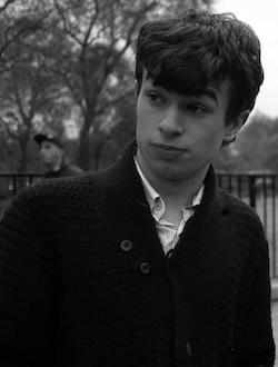 A black and white picture of a white teenage boy wearing an open-necked white shirt with a dark coloured jacket over it, standing in front of some railings behind which a man is walking; there are trees (presumably those of Hyde Park) in the background.