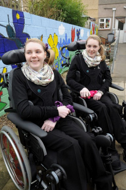 Picture of Kirstie and Catherine Fields, two young white women in black tops and black trousers sitting in wheelchairs with a wall behind them painted with flowers and butterflies.