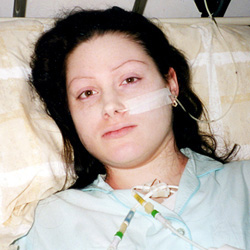 Picture of Lynn Gilderdale, a white woman with dark hair, wearing a light blue nightdress, with a naso-gastric tube and intravenous lines visible, and a pained and sad expression on her face