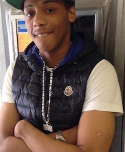 Picture of Rashan Charles, a Black teenage boy wearing a baseball cap (most of it cropped out), a short-sleeved white T-shirt with a black 'puffer' waistcoat over it. He has a long metal chain round his neck and his arms are folded.