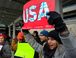 A protest at an airport, with a woman holding a sign saying 'USA: Refugees Welcome'