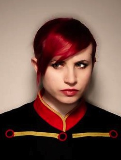 Picture of Laurie Penny, a white woman with red hair wearing a black top with a red and yellow collar and two yellow stripes across the top of the chest.