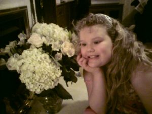 Picture of a young white girl holding her chin in her right hand, with two bundles of cream-coloured flowers next to her.