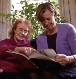 Picture of Elaine McDonald, an elderly white woman with red hair wearing a pink jumper, sitting next to a younger white woman wearing a purple jacket over a black top, holding a book at which Elaine McDonald is pointing.