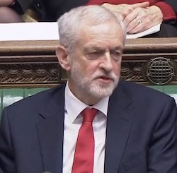 Jeremy Corbyn, an elderly white man with thin white and grey hair, sitting on the front bench of the House of Commons wearing a white shirt, red tie and dark grey suit jacket.