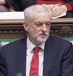 Jeremy Corbyn, an elderly white man with thin white and grey hair, sitting on the front bench of the House of Commons wearing a white shirt, red tie and dark grey suit jacket.