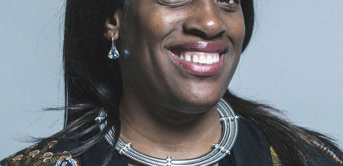 Portrait of Kate Osamor, a Black woman wearing a necklace consisting of 3 solid metal bands, with a top consisting of yellow shapes on a black background.