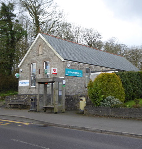 A stone building with signs saying "Pharmacy", "Post Office" and "Roche Pharmacy" on the sides. In front of it is a low wall, in front of which is a bench, a litter bin and a bus stop. The road is in the foreground.