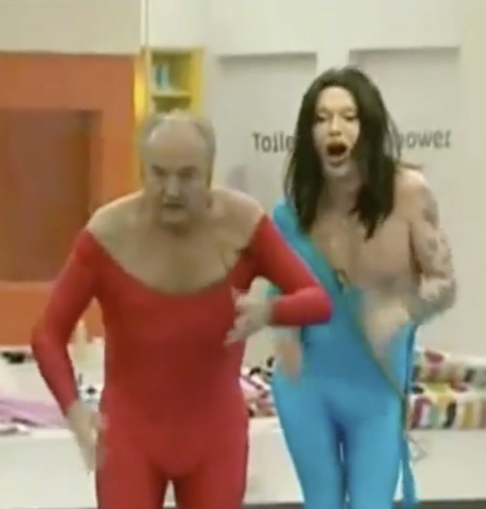 Two white men in close-fitting all-in-one bodysuits. George Galloway, the older of the two, has a red one on which has a very large scooped neckline, and Burns is (apparently) younger, has a face that looks like a woman's and has a turquoise suit which leaves his left shoulder and part of his torso bare. Two doors are behind them with "Toilet" and "Shower" on them.