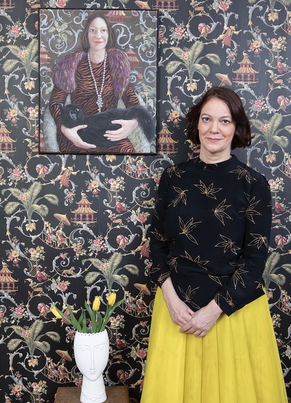 Picture of Liz Hoggard, a white woman in early middle age with brown hair wearing a bright yellow pleated skirt and a black high-necked top with black leaf or flower motifs on it, with her arms folded in front of her, in front of a wall with paper showing flowers, animals and other images on  a black background. Above her to the left is a portrait of her cradling a black cat, against the same wallpaper.