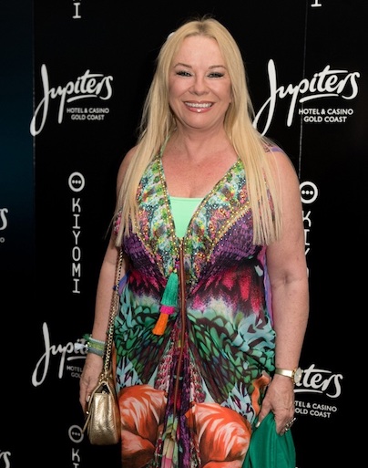 Picture of Pamela Stephenson Connolly, an older white woman wearing a dress with bright patterns in purple, green, red, orange and other colours with a gold-colour handbag over her right shoulder and holding a bottle-green scarf in her left hand. Behind her the wall has logos that read "Kiyomi" and "Jupiters hotel & casino, Gold Coast".