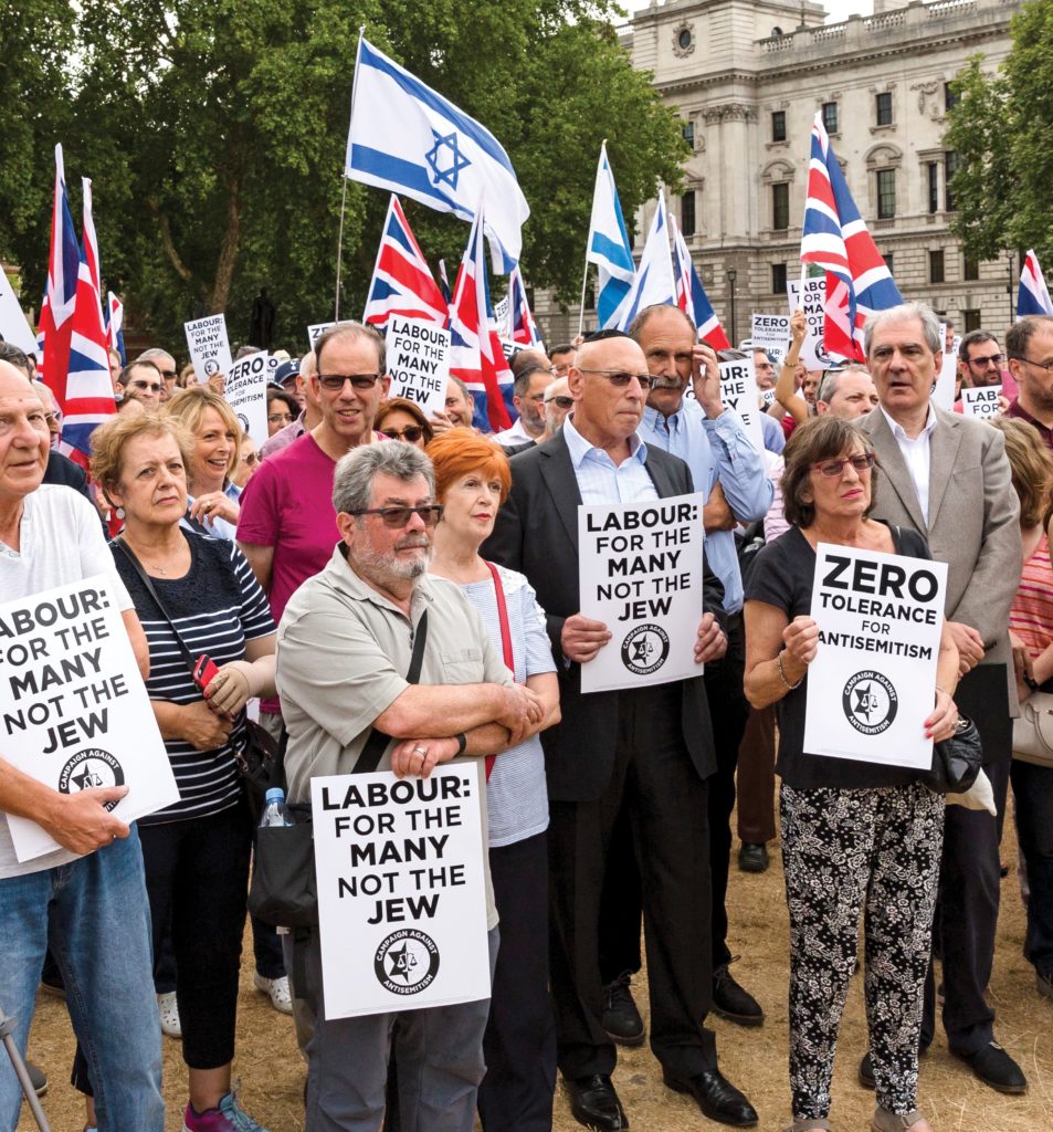 A group of people standing outside a government building in London holding British and Israeli flags with placards reading "Zero tolerance for Antisemitism" and "Labour: for the many not the Jew".