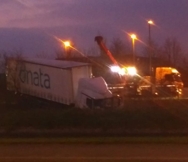 A white Mercedes articulated lorry with the "dnata" logo in blue on the side curtain. Its cab is in a stream and being pulled out by a red mobile crane during the night.