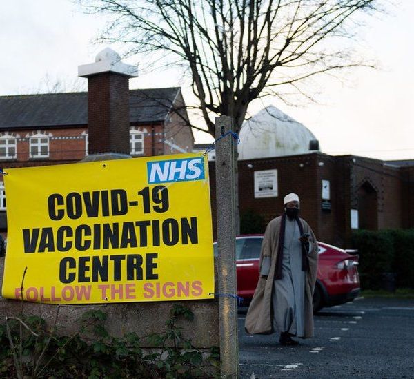 A picture of a red-brick mosque with a white dome above it with a yellow sign in the foreground reading "NHS: Covid-19 Vaccination Centre. Follow the signs". A man in a long light-blue robe and a white 'topi' hat is walking through the car park.