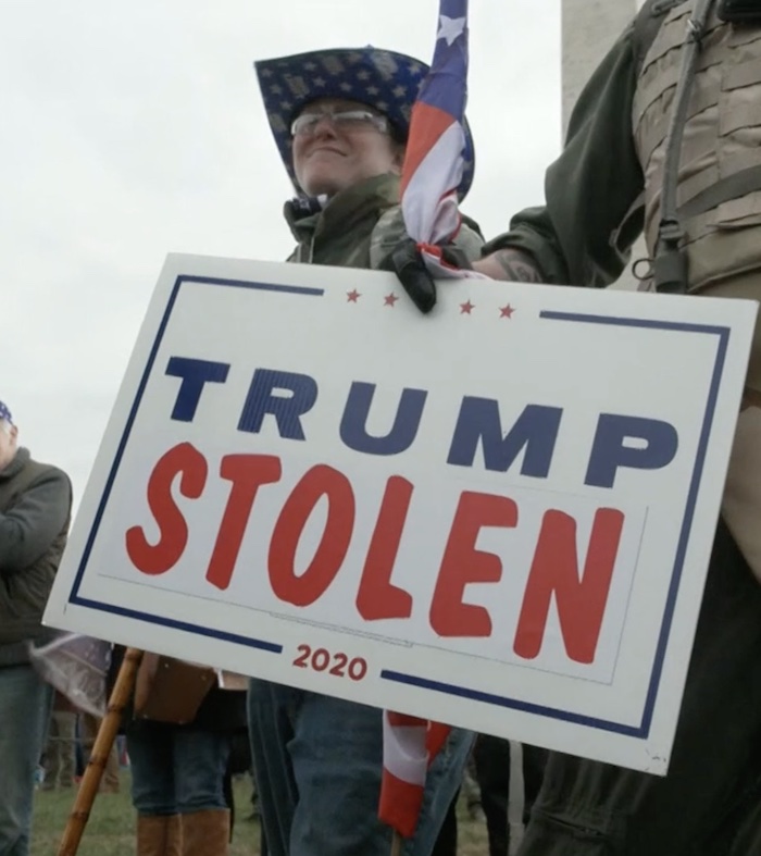 A white woman standing in a crowd next to a man in khaki clothes holding an American flag and a sign saying "Trump Stolen 2020".