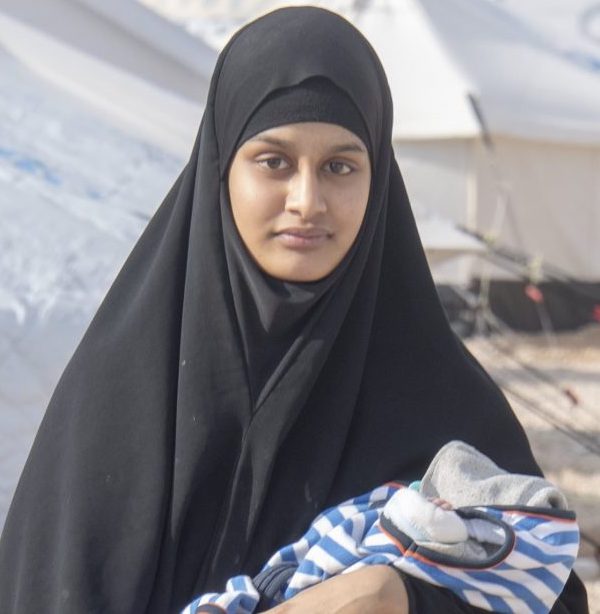 Picture of Shamima Begum, a young South Asian woman wearing a black headscarf which drapes over her body, carrying a baby who is dressed in a blue-and-white striped garment. Behind her are two large white tents.