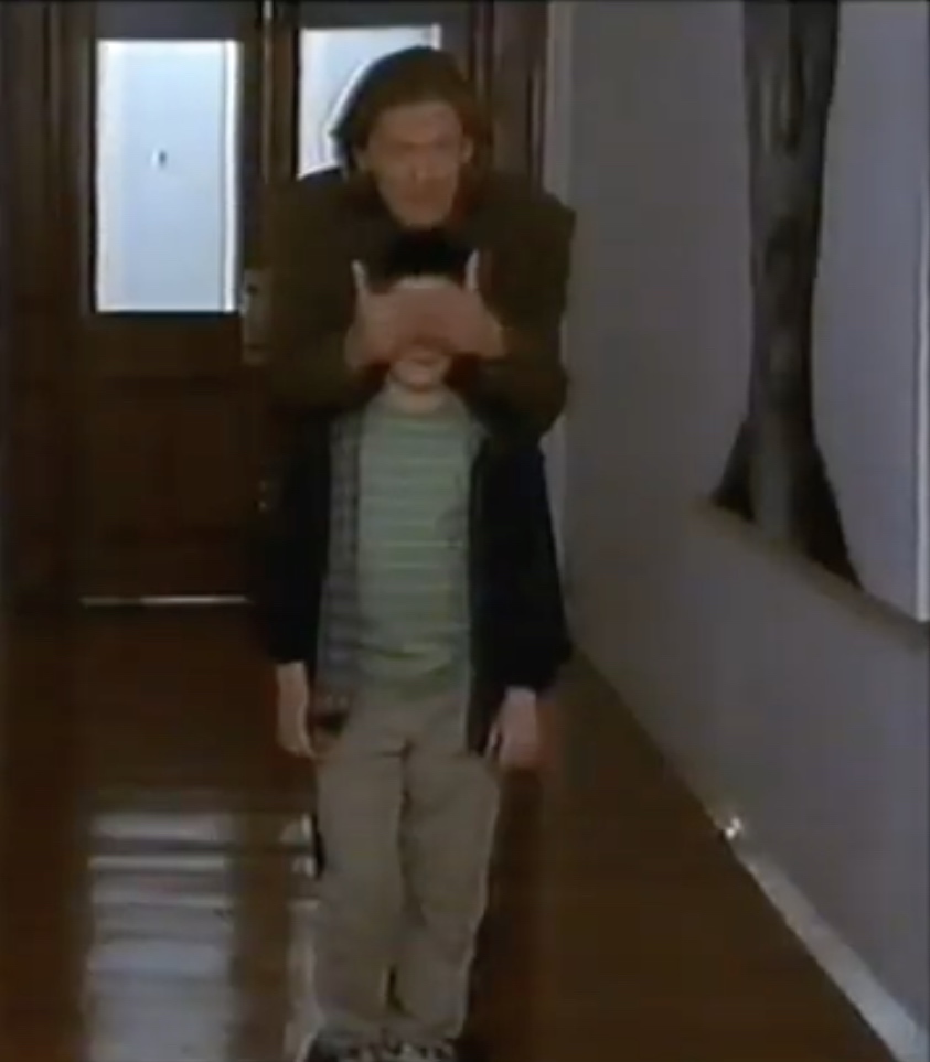 A still from a TV drama in which a man has his hands over his son's eyes in what looks like a school corridor. (He uncovers them shortly afterwards.)