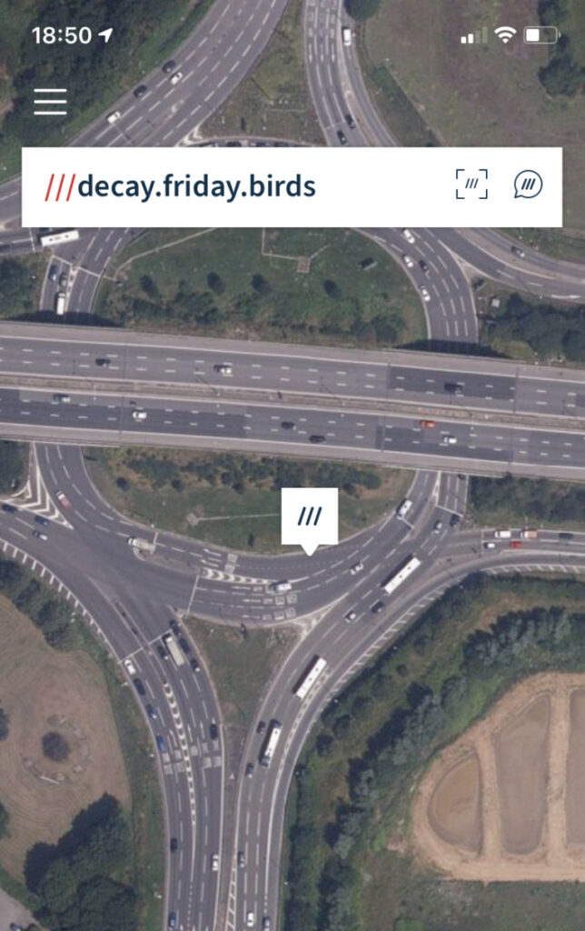A screenshot of What3Words on an iPhone showing junction 4 of the M4 (a motorway running east to west above a roundabout from which dual carriageways enter from north and south, with fields in each corner) with the W3W code "decay.friday.birds" near the top.