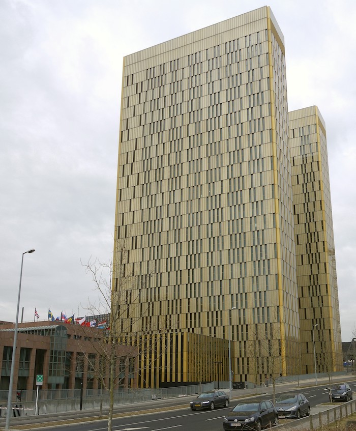 A picture of two beige skyscrapers with tall rectangular windows at random intervals. A series of different European flags can be found to the left in front of a two-storey red-brick building.