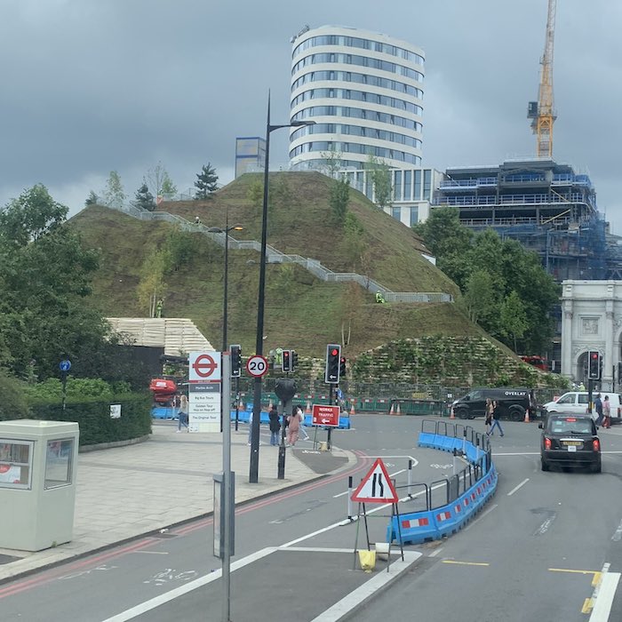A picture of a small artificial hill with thin vegetation and small trees with a metal staircase running up it. Part of Marble Arch can be seen to its right and a road, cars and a blue traffic barrier are in the foreground.