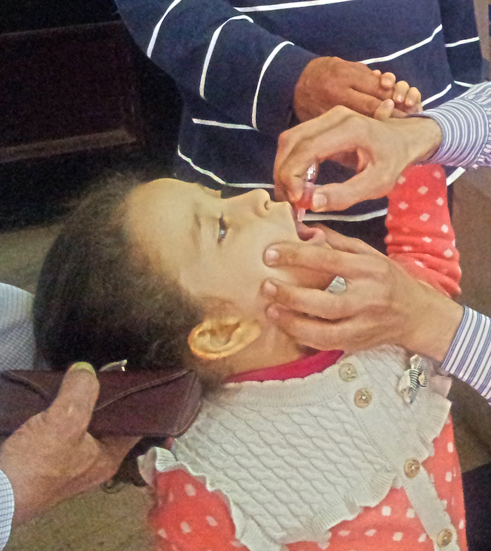 A young girl with light brown skin receiving an oral polio vaccine (a red liquid being poured into her mouth through a small vial). Two men hold her hand and support her head while a third holds her mouth open with one hand and administers the vaccine with the other.