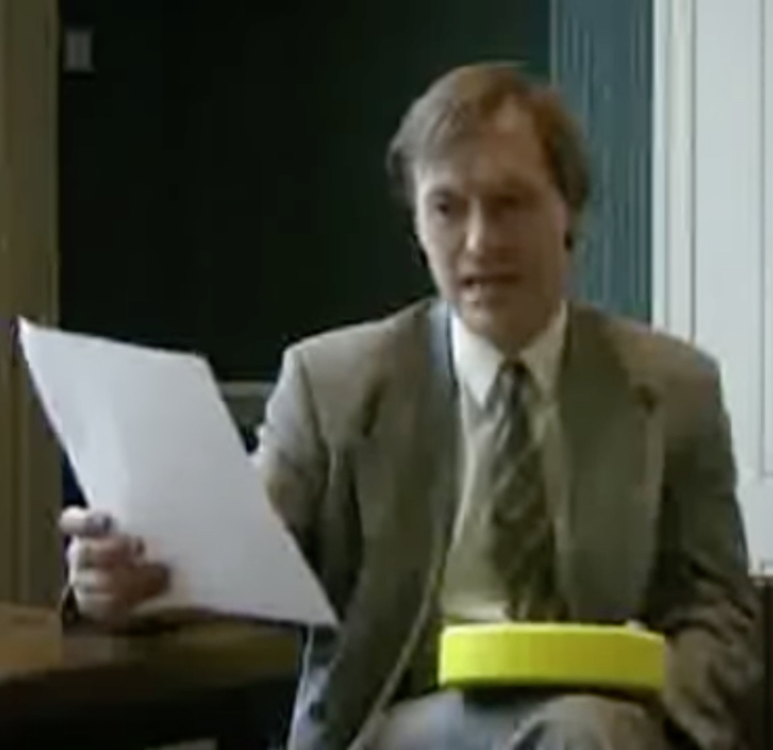 Picture of the MP David Amess in 1997, holding a yellow disc-shaped box in his left hand and reading from a piece of paper in his right hand. He is wearing a beige suit with a white shirt and a blue and yellow tie.