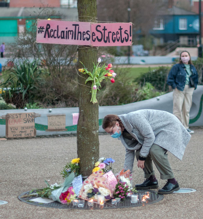 A white woman wearing a blue face mask laying flowers at the base of a tree where a number of other flowers have been left. A banner attached to he tree reads "Reclaim these streets" and other banners can be seen on the wall behind, including one saying "Rest in love Sarah; you deserved to make it home safe". Another white woman, wearing a pink mask, if standing in front of the wall.