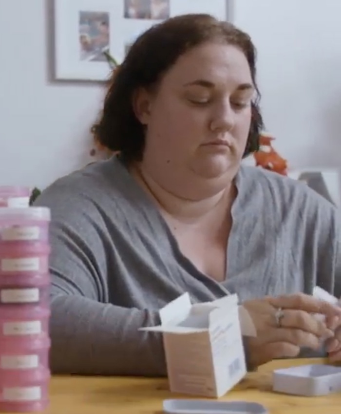 Picture of Lucy Wilson, a white woman in her 30s wearing a grey T-shirt, sitting at a wooden table with boxes of tablets in front of her
