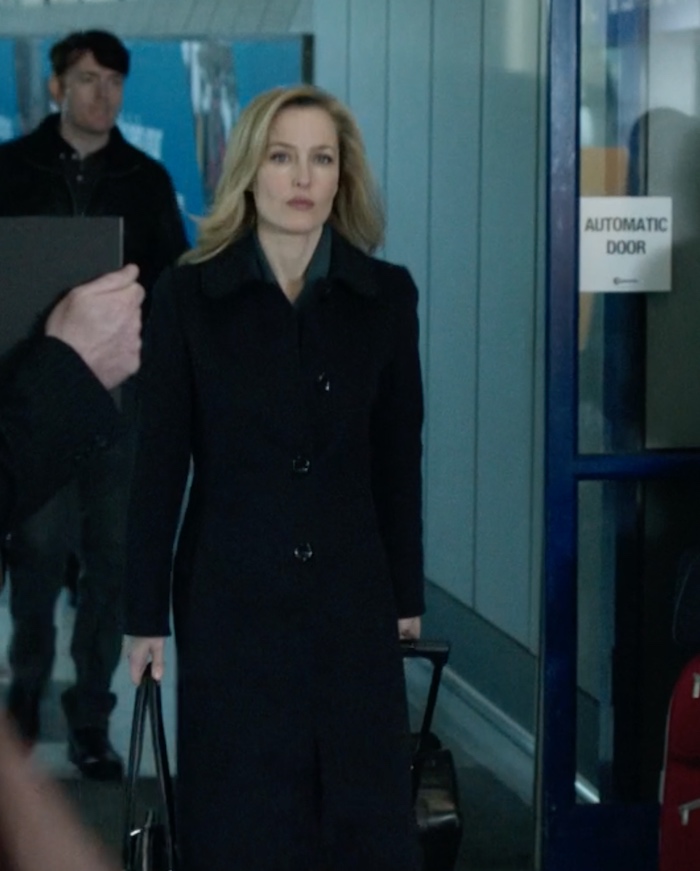 Picture of a middle-aged white woman in a black suit with an above-knee skirt, walking through an airport pulling a suitcase behind her and carrying another bag in her right hand.