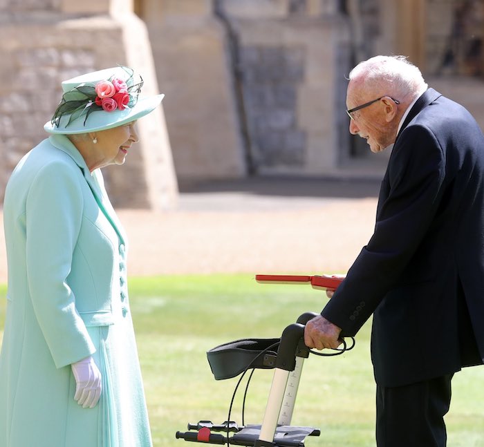 The Queen, an elderly white woman wearing a light turquoise coat and a hat of the same colour with roses mounted on it, facing Captain Sir Tom Moore, an elderly white man wearing a dark grey suit, with one hand on his walker and holding the red box containing his knight's medal in his right hand. The walls of a castle (Windsor) are blurred behind them.