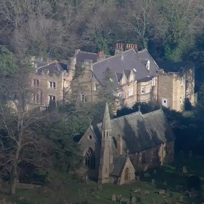 An aerial picture of a large stone country house with a tall roof in a wood, with a church and steeple in the foreground.