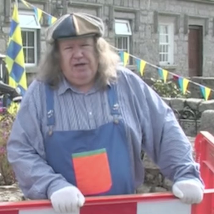 Picture of Richie Kavanagh, an older white man with long, curly fair hair wearing blue dungarees over a white and blue stuffed shirt, standing behind a red plastic barrier of the sort used to fence off works on pavements. Behind him is a stone house festooned with blue and yellow flags.