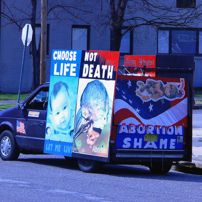 A picture of a blue van with its rear covered by large pictures of babies and foetuses with the slogans "Choose life not death", "Let me live", "Abortion shame", "Abortion causes breast cancer" and "Hitler's Holocaust" printed on them. There is an American flag transfer on the left side of the van.