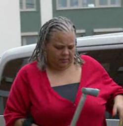 A light brown-skinned woman with braided hair, wearing a red jumper and a black top under it, holding a crutch in her hand, getting out of a metallic grey Nissan car