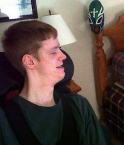 Picture of Hunter Dunn, a young white man with cerebral palsy, wearing a green jumper, sitting in a wheelchair looking to his left (at an off-picture computer screen)