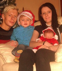 Picture of a young white man and woman sitting on a sofa with a toddler and baby son, with a Christmas tree on the left