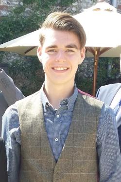 Picture of Oliver McGowan, a young white man with light brown hair combed from a parting on the left side, wearing a blue and white patterned shirt and a tweed waistcoat, standing in front of a large parasol.