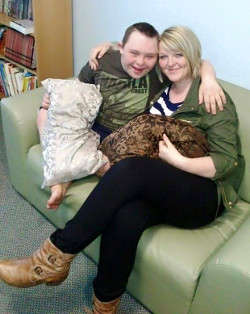 A picture of Thomas Rawnsley, a young man with Down's syndrome wearing a green/khaki T-shirt, sitting on a sofa next to Becky, a young woman with blonde hair wearing a blue and white striped T-shirt, a green jacket, black leggings and brown leather boots.