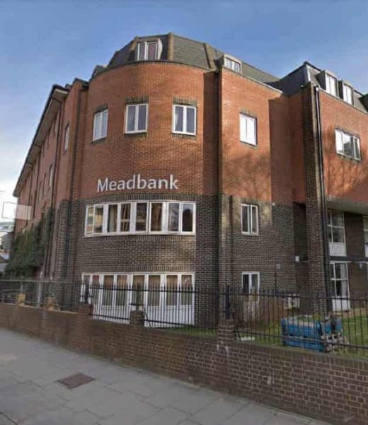 A picture of Meadbank care home, a red-brick building on a corner with the name 'Meadbank' in large white letters on the rounded corner, with a brick wall topped by a black metal railing in front of it.