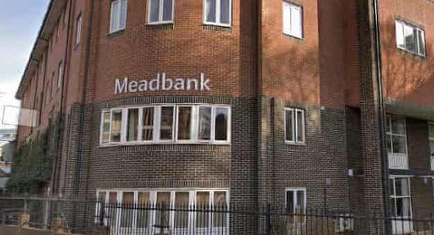 A picture of Meadbank care home, a red-brick building on a corner with the name 'Meadbank' in large white letters on the rounded corner, with a brick wall topped by a black metal railing in front of it.