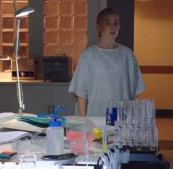 Still from Holby City of Jac Naylor, a middle-aged white woman wearing no make-up and with blonde hair tied behind her head, wearing a hospital gown, standing in a darkened laboratory in front of a table with a desk lamp shining down and some test tubes in a rack.
