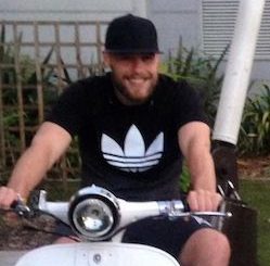 Dale Hart, a white man wearing a black cap and shorts and a black T-shirt with a white Adidas logo, sitting on a white scooter.