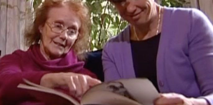 Picture of Elaine McDonald, an elderly white woman with red hair wearing a pink jumper, sitting next to a younger white woman wearing a purple jacket over a black top, holding a book at which Elaine McDonald is pointing.