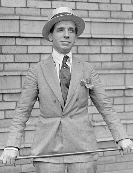 Black and white picture of Charles Ponzi, a white man dressed in a suit and tie with a wide-rimmed hat, holding a walking stick in his hands.