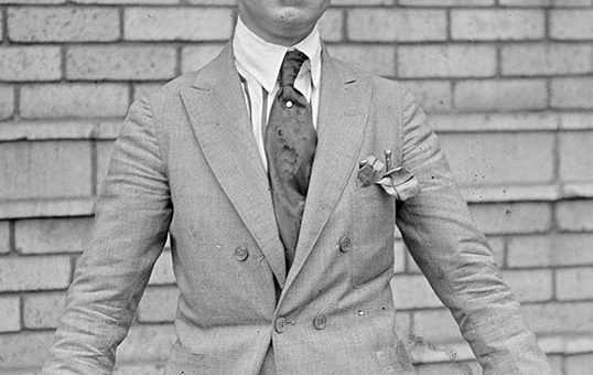 Black and white picture of Charles Ponzi, a white man dressed in a suit and tie with a wide-rimmed hat, holding a walking stick in his hands.