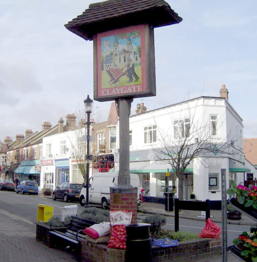 Picture of a village sign showing a church and a man pushing a loaded barrow across grass in front of it; the sign stands on a pole amid a flowerbed surrounded by benches and a roadsign facing the other way. There is a parade of shops across the road which stretches from a corner behind the sign, including a florist with flowers displayed outside at the shop on the corner.